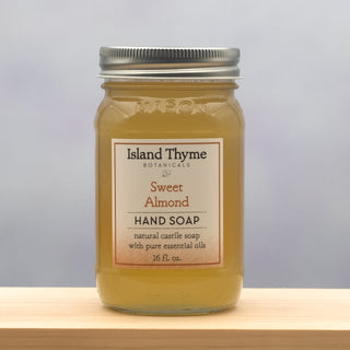 A mason jar labeled Sweet Almond Castile Soap sits on a wooden surface, filled with light amber liquid soap. The label notes it contains natural castile soap with pure essential oils, 16 fl. oz.