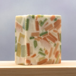 Confetti Soap features a white rectangular base with colorful orange, green, and yellow rectangular pieces embedded inside, shown on a light wooden surface with a soft gradient background.