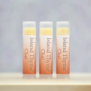 Three tubes of Chai Lip Balm stand upright on a light surface against a soft purple background. Their labels are clearly visible, featuring the product name and highlighting its gluten-free formula.