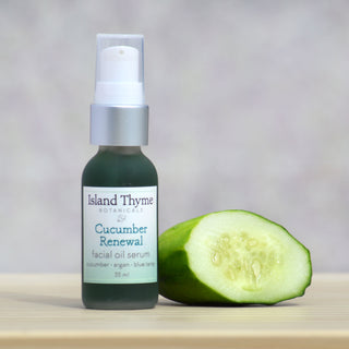 A bottle of Cucumber Renewal Oil Serum rests beside a halved cucumber on a light wooden surface, emphasizing its nourishing and anti-inflammatory qualities against a softly blurred background.