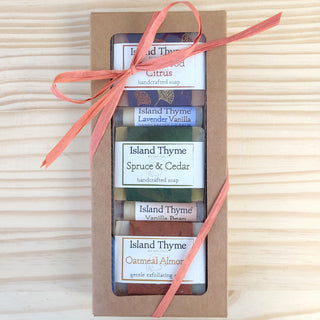 The Earthy Gift Box features five handcrafted Island Thyme soaps, each with a unique scent—including woody forest notes—individually labeled and tied with a pink ribbon, displayed on a light wood surface.