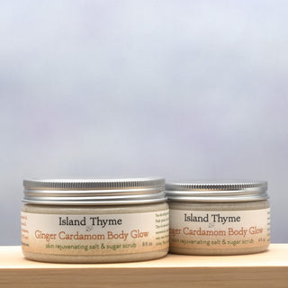 Two jars of Ginger Cardamom Body Glow Salt & Sugar Scrub, one large and one small, rest on a wooden surface against a blue-gray background—ready to exfoliate and moisturize your skin for a smooth, radiant glow.