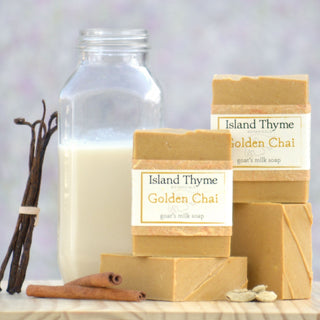 A glass bottle of milk, vanilla beans, cinnamon sticks, and cardamom pods rest next to stacked bars of Golden Chai Goat's Milk Soap on a wooden surface with a pale background.