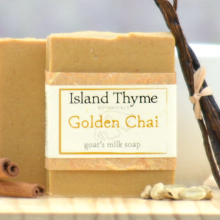 A handmade Golden Chai Goat's Milk Soap bar stands upright beside another, with cinnamon sticks, cardamom pods, and a vanilla bean in the background.