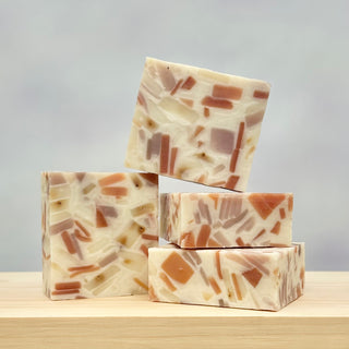 Four handcrafted Confetti Soap bars with terrazzo patterns in cream, pink, and brown are stacked on a light wood surface against a softly blurred background.