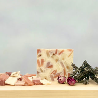 A bar of Confetti Soap stands on a wooden surface, surrounded by colorful soap pieces and dried flowers, including purple sprigs and pink rosebuds, all set against a soft light blue background.