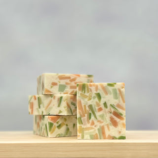 Four rectangular Confetti Soap bars, with embedded green, orange, and cream pastel fragments, are stacked on a light wooden surface. Handmade for beauty and sustainability, each bar helps reduce waste—set against a softly blurred background.