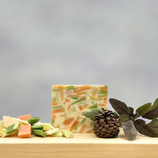 The Confetti Soap, featuring vibrant colorful chunks, is displayed with scattered soap pieces, a pine cone, and green leaves on a wooden surface against a soft neutral background.