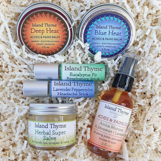 The Remedies & Soothers Deluxe set, featuring Island Thyme’s natural balms, salves, and sticks, including Deep Heat, Blue Heat tins, a headache stick roll-on, herbal salve jar, and Aches & Pains oil bottle, displayed on shredded paper.