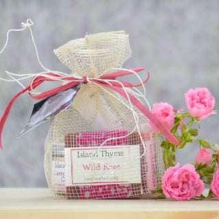 The Rose Gift Set features handcrafted Wild Rose soap by Island Thyme in a burlap bag with a red ribbon, accented by pink roses, all arranged on a light wooden surface against a softly blurred background.