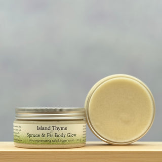 A jar of Spruce & Fir Body Glow Salt & Sugar Scrub rests on a light wooden surface—one jar upright with the label visible, and another open to display the exfoliating scrub inside.