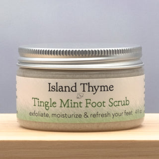 A 4 fl oz jar of Tingle Mint Foot Scrub with a silver lid sits on a wooden surface. The label highlights its exfoliating, moisturizing, and refreshing benefits for feet.