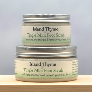 Two containers of Tingle Mint Foot Scrub are stacked on a wooden surface, their labels showing this refreshing and exfoliating mint foot scrub. The light background appears softly blurred.
