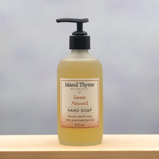 A bottle of Sweet Almond Castile Soap with a pump dispenser sits on a light wooden surface against a neutral background. The 8 fl. oz. label highlights pure essential oils.
