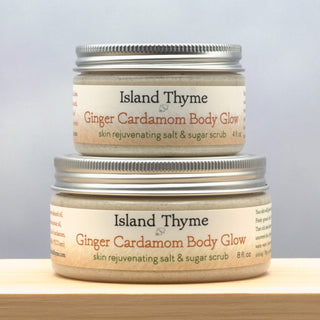 Two jars of Ginger Cardamom Body Glow Salt & Sugar Scrub, one 4 oz. and one 8 oz., are stacked on a wooden surface against a soft gray background, showcasing this exfoliating scrub for radiant skin.