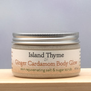 A jar of Ginger Cardamom Body Glow Salt & Sugar Scrub, with an exfoliating formula for smooth, moisturized skin, rests on a wooden surface. Its silver lid and beige-white label with green and orange text enhance its fresh look.