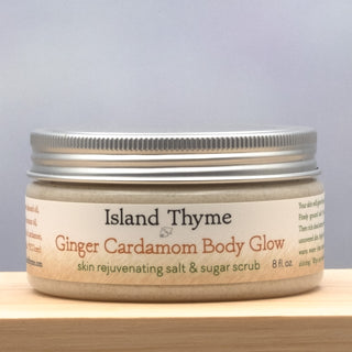 A jar of Ginger Cardamom Body Glow Salt & Sugar Scrub, an exfoliating formula for smooth, moisturized skin, features a cream-colored label with brown and green text and a silver lid on a wooden surface.