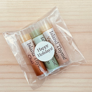 A clear plastic bag holds the Lip Balm - Holiday Sampler 3-Pack by Island Thyme, featuring Chai, Mint, and Vanilla Bean flavors. A round “Happy Holidays!” sticker decorates the natural lip balm set on a light wood surface.