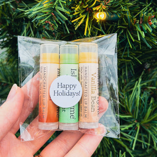 A hand holds the Lip Balm - Holiday Sampler 3-Pack, featuring three colorful Island Thyme lip balms labeled “Happy Holidays!”—the perfect stocking stuffer, displayed in front of a festive Christmas tree.