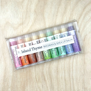 A clear plastic package containing nine tubes of Island Thyme Lip Balm—natural and gluten-free—displayed on a light wood surface. Each tube in the Lip Balm - Complete Set of 9 is labeled with its unique flavor.
