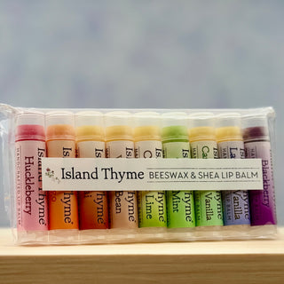 The Lip Balm - Complete Set of 9 comes in a clear bag with nine natural, gluten-free Island Thyme lip balms in flavors like Huckleberry, Cinnamon, Honey, Bean, Lime, Vanilla, and Blackberry, all lined up neatly.