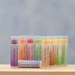 A complete set of 9 Island Thyme Lip Balms, all natural and gluten-free, in flavors like Blackberry, Mango, and Tangerine Mint, is arranged in a row on a wooden surface.