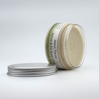 A clear jar of Spruce & Fir Body Glow Salt & Sugar Scrub with a white lid beside it. The creamy exfoliating scrub is visible inside, and the jar features green and white branding.
