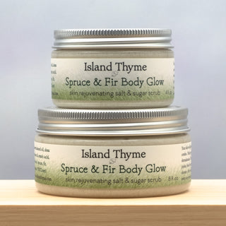 Two jars of Spruce & Fir Body Glow Salt & Sugar Scrub are stacked on a wooden surface, with the larger jar on the bottom and the smaller one neatly placed on top.