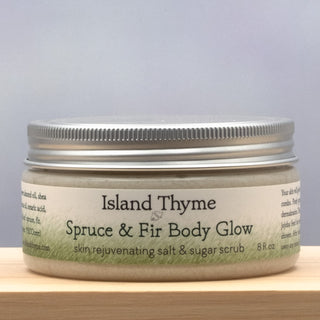 An 8-ounce jar of Spruce & Fir Body Glow Salt & Sugar Scrub, an exfoliating scrub with a silver lid, rests on a light wooden surface against a pale background.