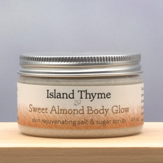 A 4 fl. oz. jar of Sweet Almond Body Glow Salt & Sugar Scrub, an exfoliating and moisturizing body scrub, rests on a light wooden surface with a neutral background. The label showcases earthy tones and clear text.