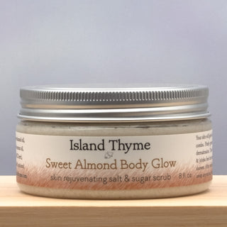 A jar of Sweet Almond Body Glow Salt & Sugar Scrub features a silver lid, cream-colored label, and contains 8 fl. oz. of exfoliating, moisturizing body scrub.