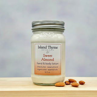 A glass mason jar labeled Sweet Almond Lotion rests on a wooden surface beside whole almonds, showcasing this moisturizing lotion with an invigorating scent against a softly blurred gradient background.
