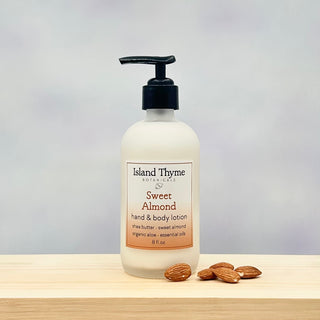 A bottle of Sweet Almond Lotion with a black pump rests on a light wooden surface beside whole almonds, set against a soft gray background.