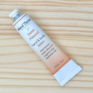 A travel-size tube of Sweet Almond Lotion with shea butter, sweet almond, organic aloe, and essential oils featuring an invigorating scent rests on a light wood surface.