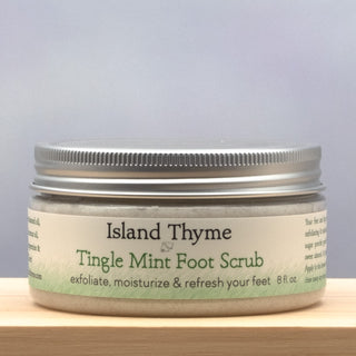 An 8 fl oz jar of Tingle Mint Foot Scrub with a silver lid, labeled as an exfoliating scrub to moisturize and refresh feet, sits on a wooden surface against a neutral background.