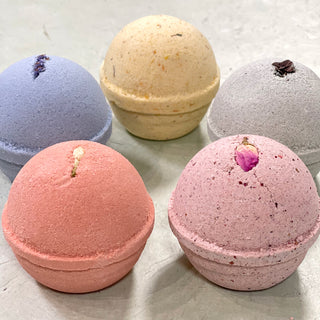 The Bath Bomb Sampler 5 Pack features pink, blue, yellow, gray, and peach bath bombs, each topped with a dried flower or bud and arranged on a light gray surface—perfect as an essential oil bath bomb gift set.