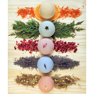 The Bath Bomb Sampler 5 Pack features five pastel bath bombs arranged vertically on wood, each above its matching natural ingredient: orange and carrot, evergreen branches, rose petals, lavender buds, and mixed spices.