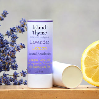 A tube of Lavender Lemon Deodorant stands upright beside a halved lemon and lavender flowers, with another tube lying horizontally in the background.