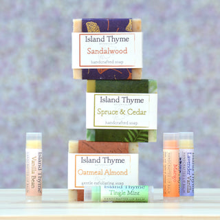 The Earthy Gift Box includes three Island Thyme handcrafted forest-scented soaps stacked vertically, paired with four scented lip balms—Vanilla Bean, Tingle Mint, Mango, and Lavender Vanilla—displayed beside them.