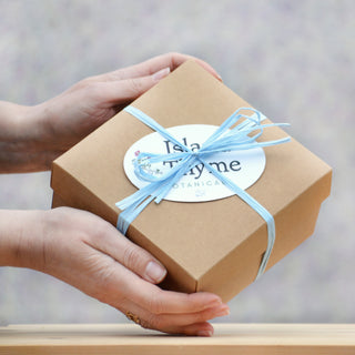 Two hands hold a brown gift box tied with a light blue ribbon and topped with a round white label. The softly blurred background evokes the gentle care of the Soothing Flowers Complete Set.