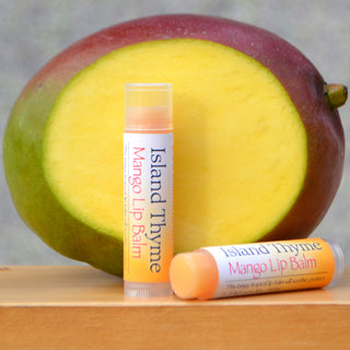 Two tubes of Mango Lip Balm, made with natural ingredients, are shown on a wooden surface in front of a sliced mango—one tube stands upright while the other lies down.