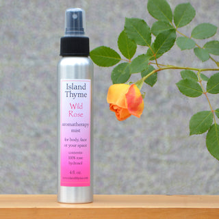 A silver 4oz spray bottle labeled Wild Rose Mist, made with rose distillate water for skin toning, is shown beside an orange-yellow rose and green leaves against a soft gray background.