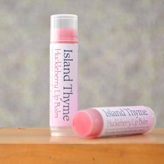 Two tubes of Huckleberry Lip Balm rest on wood—one upright, one lying down—to showcase their pink hue and white labels. Experience natural protection and deep moisturizing care. The softly blurred background enhances their appeal.