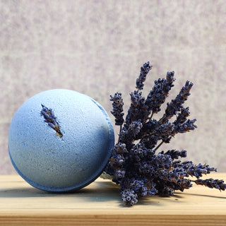 A natural bath bomb from the Bath Bomb Sampler 5 Pack sits on wood beside dried lavender, with a lavender-toned background—an ideal choice to enhance any bath bomb gift set.