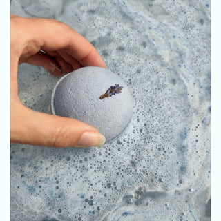 A hand holds a round, light purple bath bomb from the Bath Bomb Sampler 5 Pack, topped with dried lavender, above foamy bath water tinted light blue.