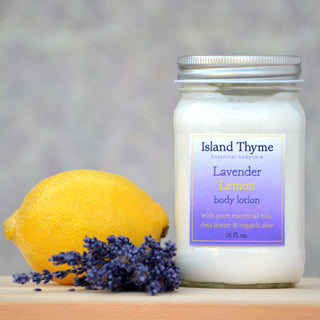 A jar of Lavender Lemon Lotion sits beside a fresh lemon and dried lavender on a light surface, with a blurred gray and purple background.