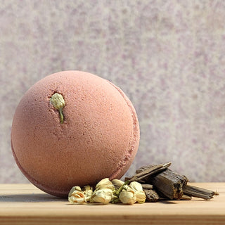 A Bath Bomb Sampler 5 Pack bath bomb, pink with a dried white flower on top, sits on wood surrounded by dried flowers and brown sticks, set against a soft speckled purple background.