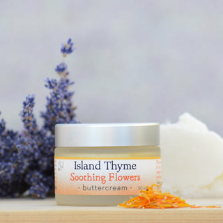 A jar of Soothing Flowers Buttercream, famed for its deep moisturizing properties, rests on wood among dried lavender and flower petals. The pastel label and silver lid add charm to this nourishing cream.