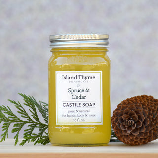 A reusable mason jar of Spruce & Cedar Castile Soap sits beside a pinecone and spruce branch against a soft, light purple background.