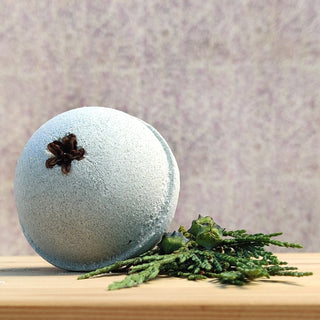 A round, pale blue bath bomb from the Bath Bomb Sampler 5 Pack, topped with star anise, rests on wood beside green foliage, set against a softly blurred purple background.
